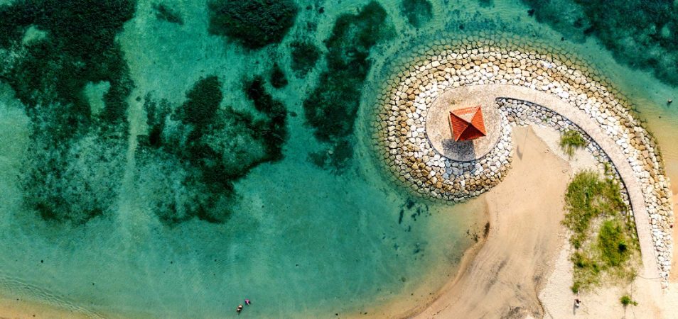 A bird's-eye view of a spiraling stone walkway on a beach by a crystal blue ocean.