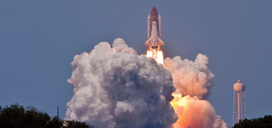 A space shuttle launches from Cape Canaveral on the Florida coast with a large plume of smoke.