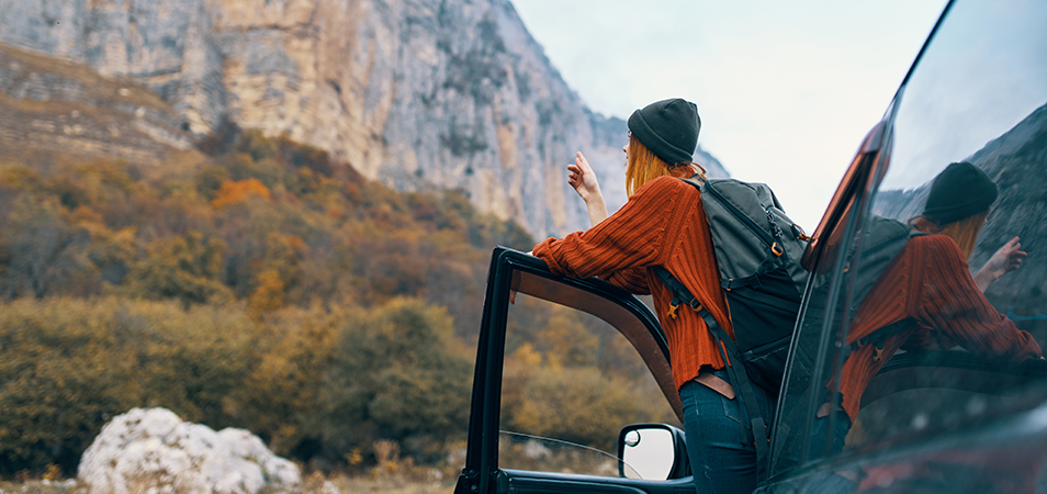 A woman leaning out of her car, pointing at a nearby mountain with trees cascading down.