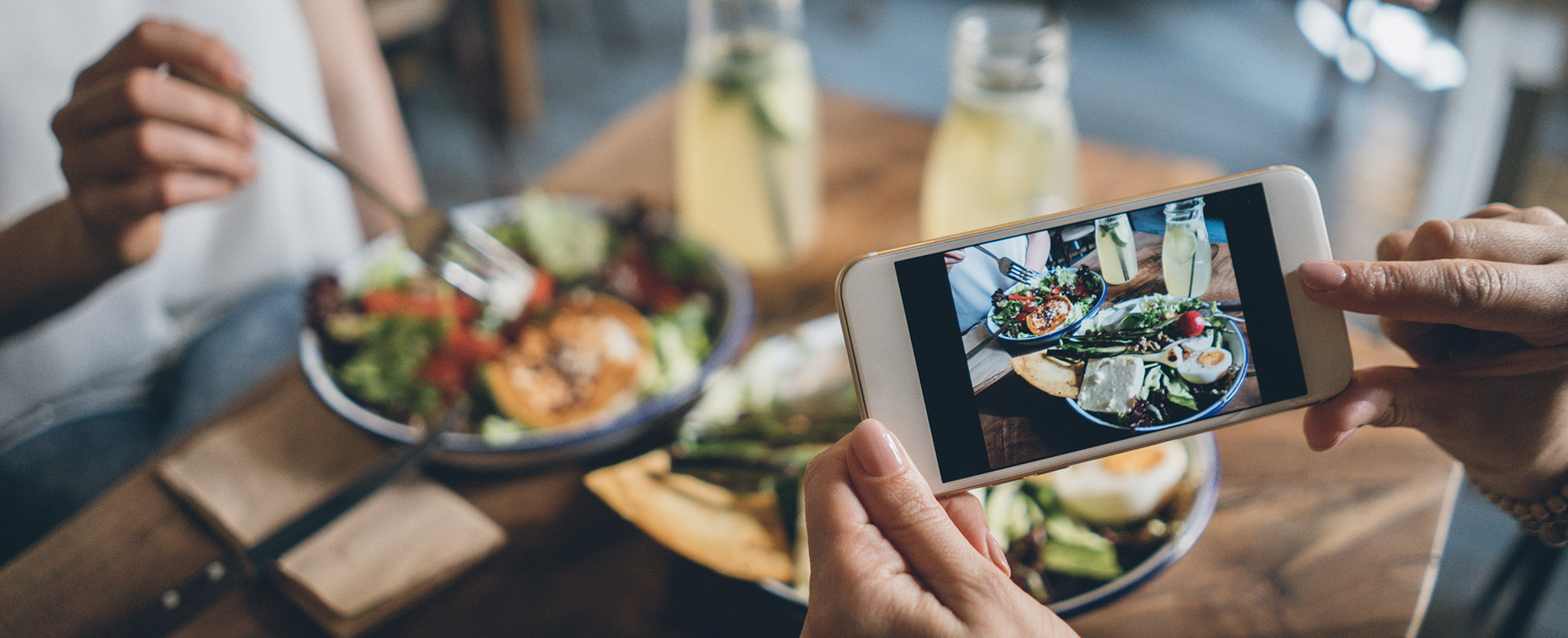 Woman holds her phone above a table for two to take a photo of their meal on a wooden table, including two full plates of food and two glass jars of lemonade.