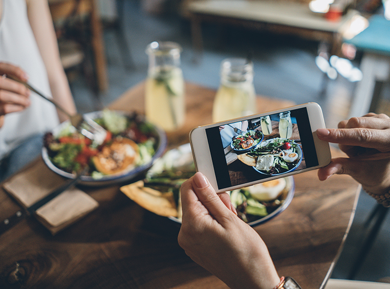 Woman holds her phone above a table for two to take a photo of their meal on a wooden table, including two full plates of food and two glass jars of lemonade.