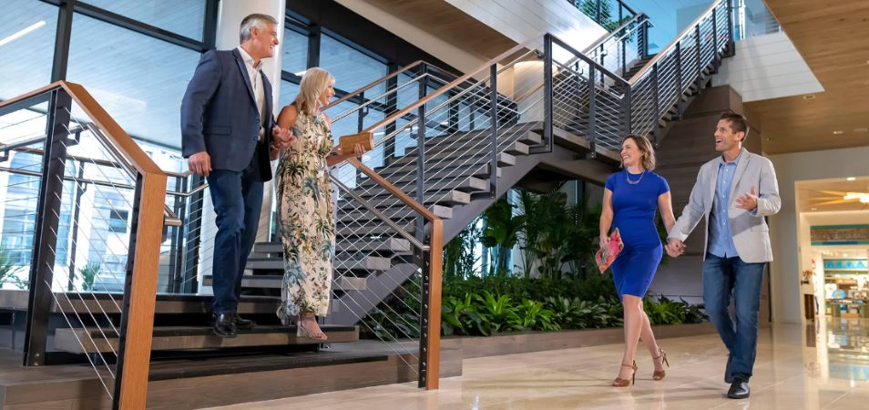 Two couples, each holding hands, greet each other at the bottom of a staircase at a Margaritaville Vacation Club resort.