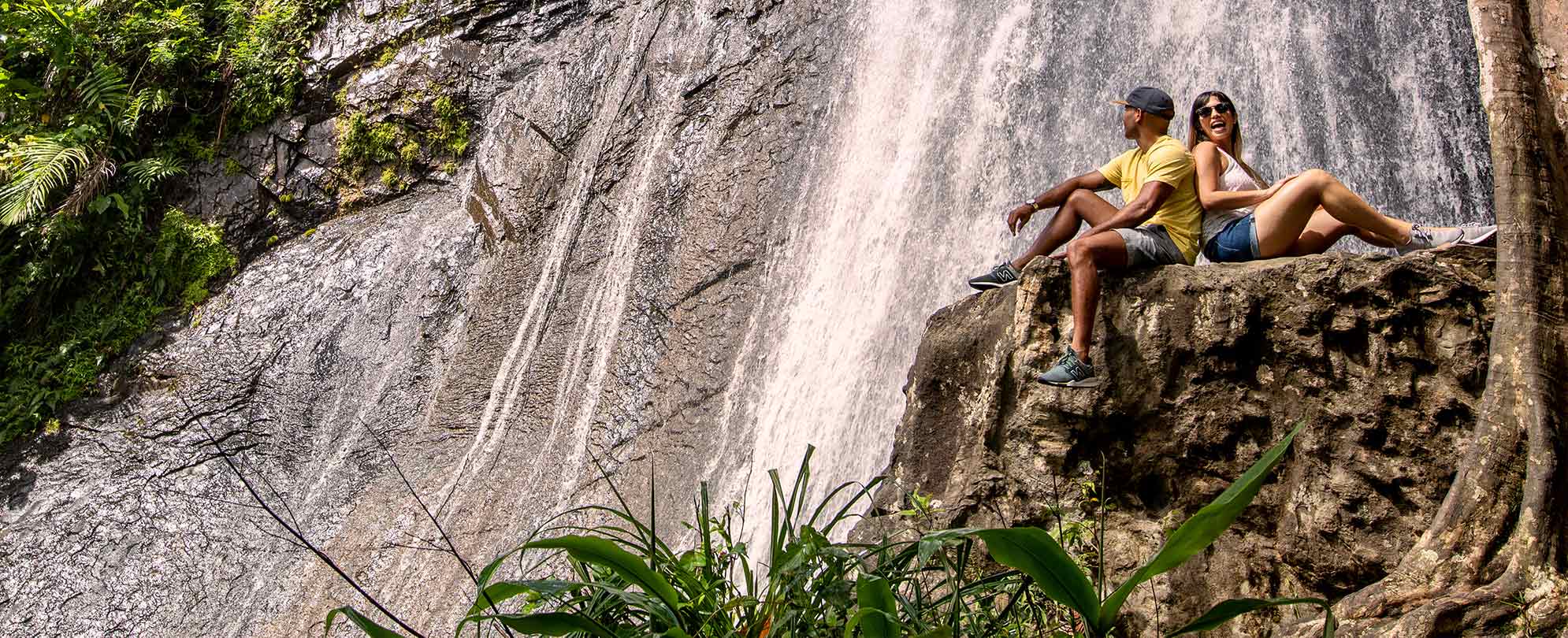 A man and woman sitting back to back on top of a large rock formation in front of a waterfall. 