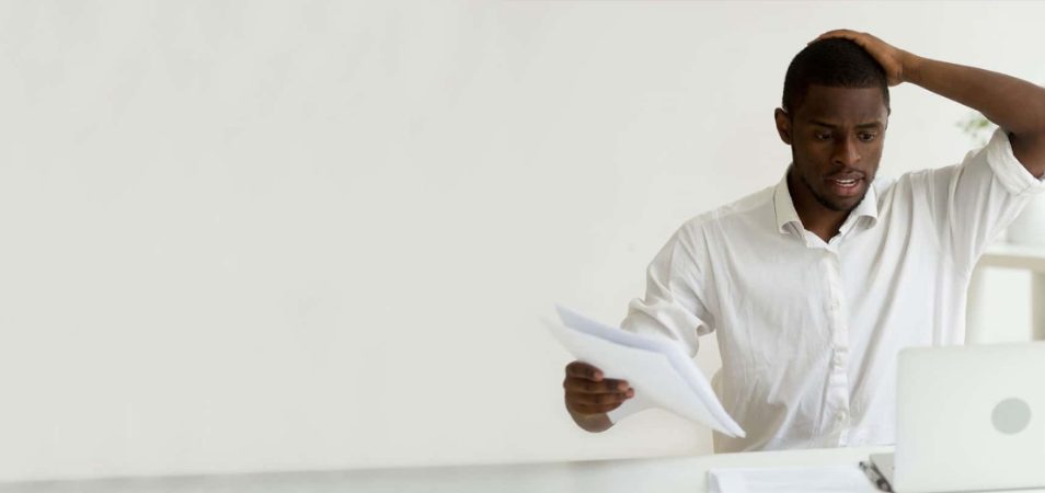 A man looking overwhelmed holding his head in one hand and paperwork in another as he sits at a desk in front of his laptop. 