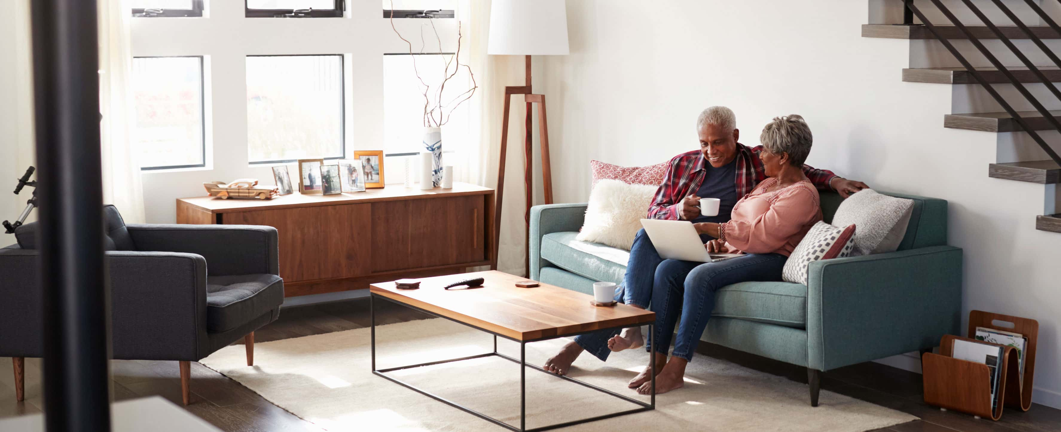 A woman with a laptop on her lap sitting close to her husband on the couch conversing with him as he holds his coffee cup. 