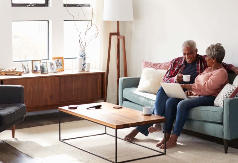 A woman with a laptop on her lap sitting close to her husband on the couch conversing with him as he holds his coffee cup. 