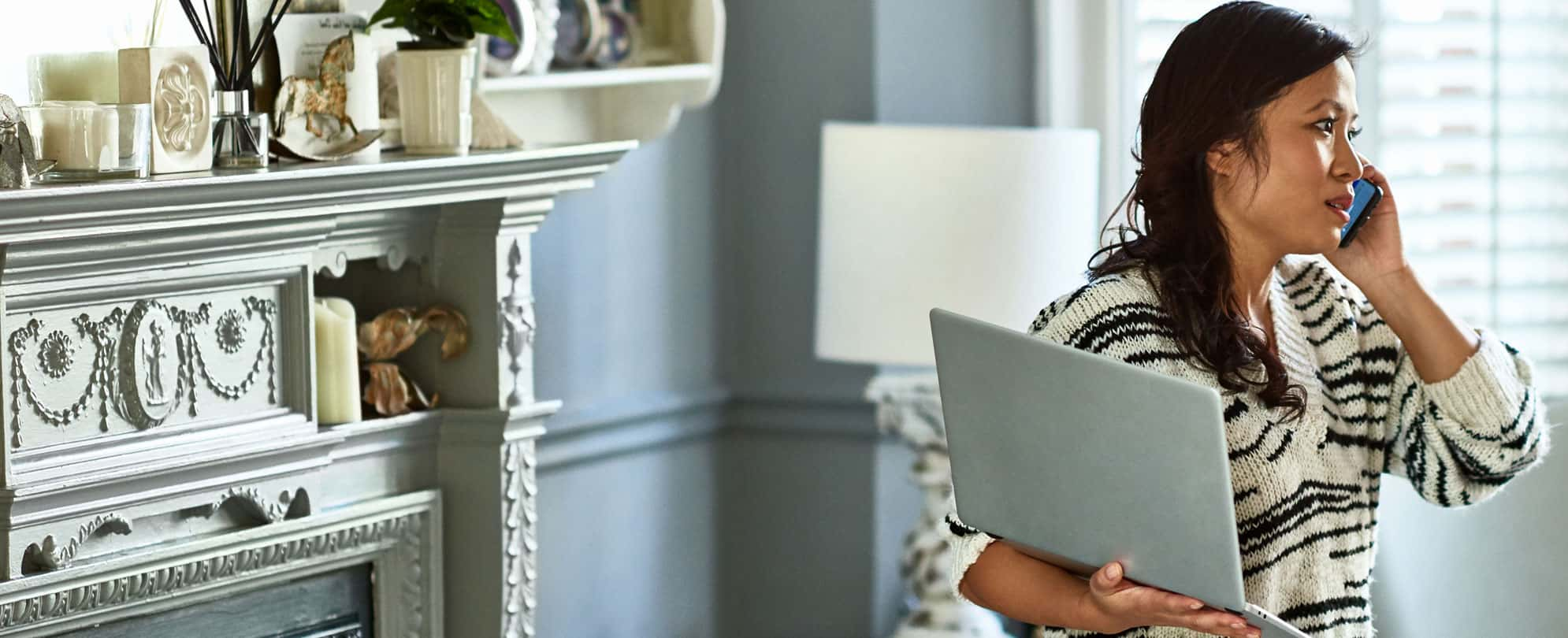 A woman in her living room holding a laptop as she has a conversation on the phone. 