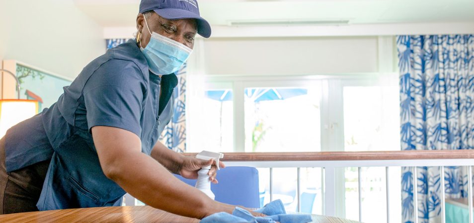 Female employee wearing blue medical mask sprays and wipes down table in a Margaritaville Vacation Club suite.