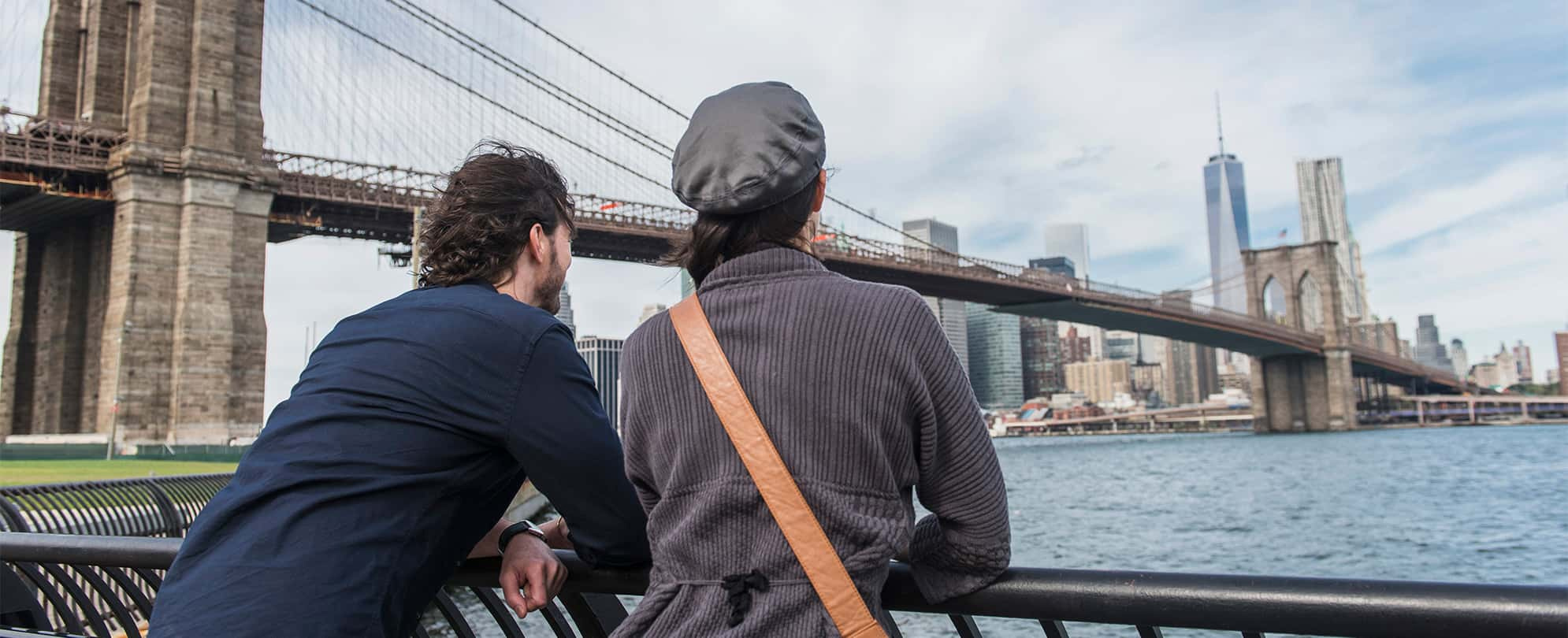 Man and woman leaned over railing, taking in the view of the New York City skyline and Brooklyn Bridge.