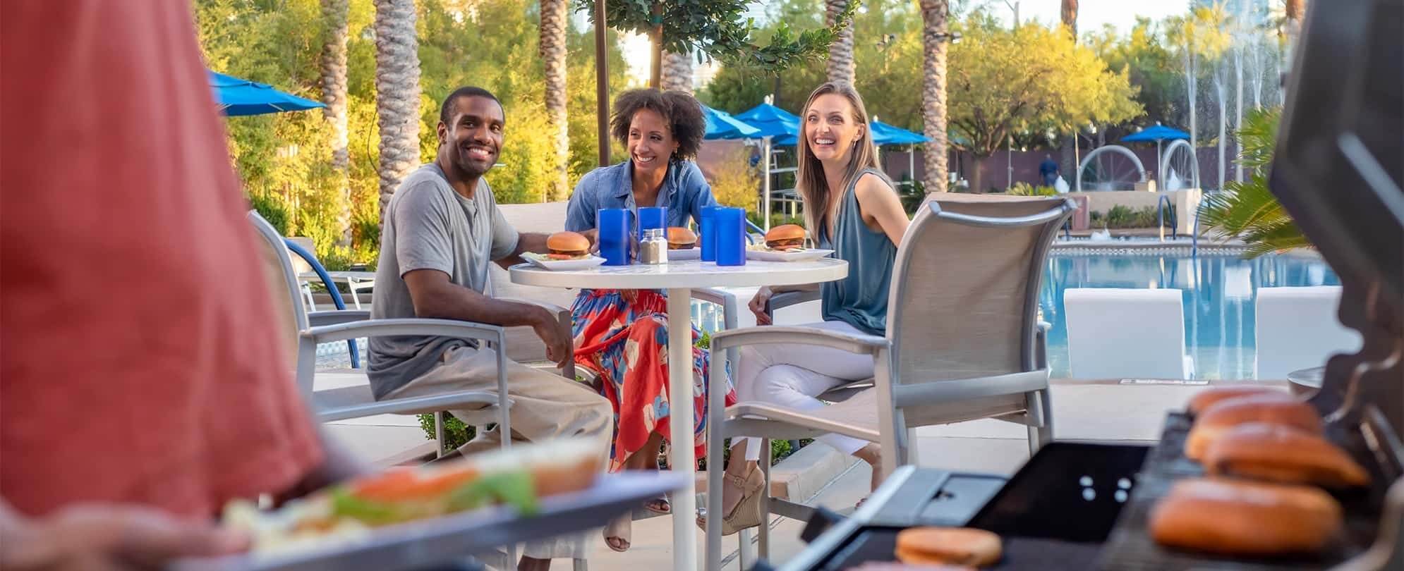 Three adults sitting at a poolside table enjoy grilled burgers at a Margaritaville Vacation Club resort.