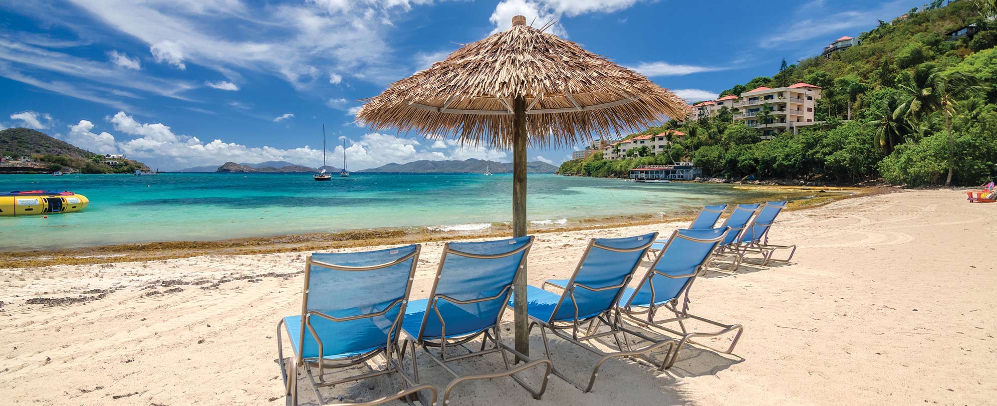 Blue beach chairs sit under a straw umbrella in front of the ocean at Margaritaville Vacation Club by Wyndham - St. Thomas.