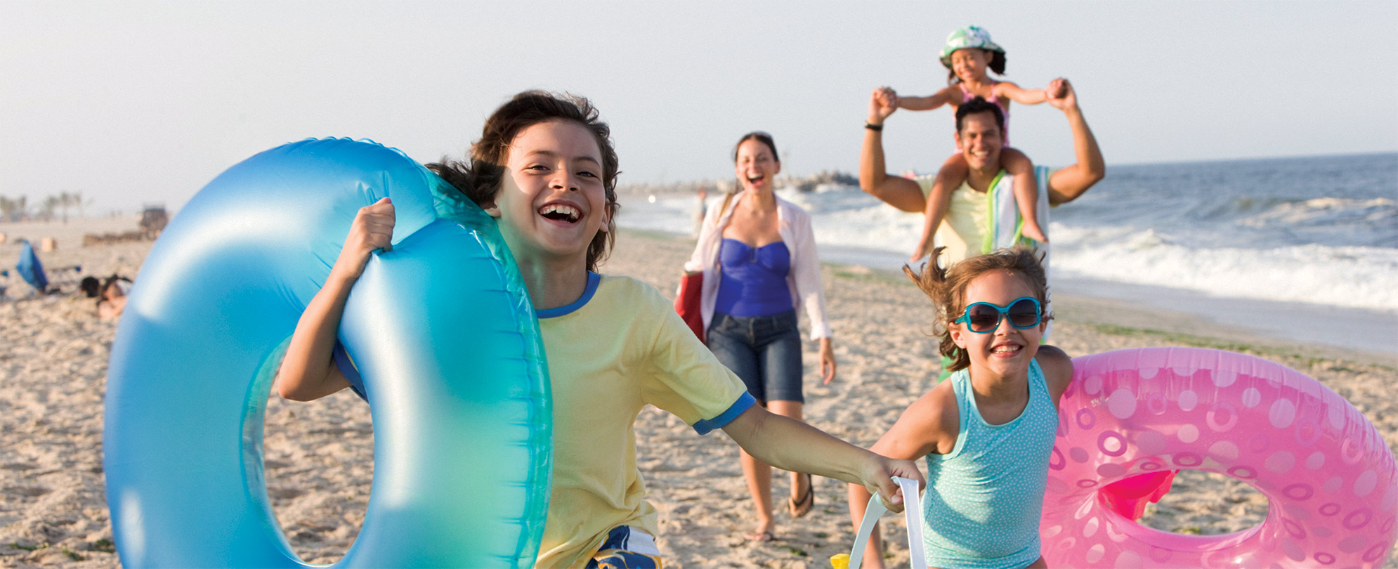 Two kids carrying inflatable tubes walk in front of their mom, dad, and sibling along a national seashore.