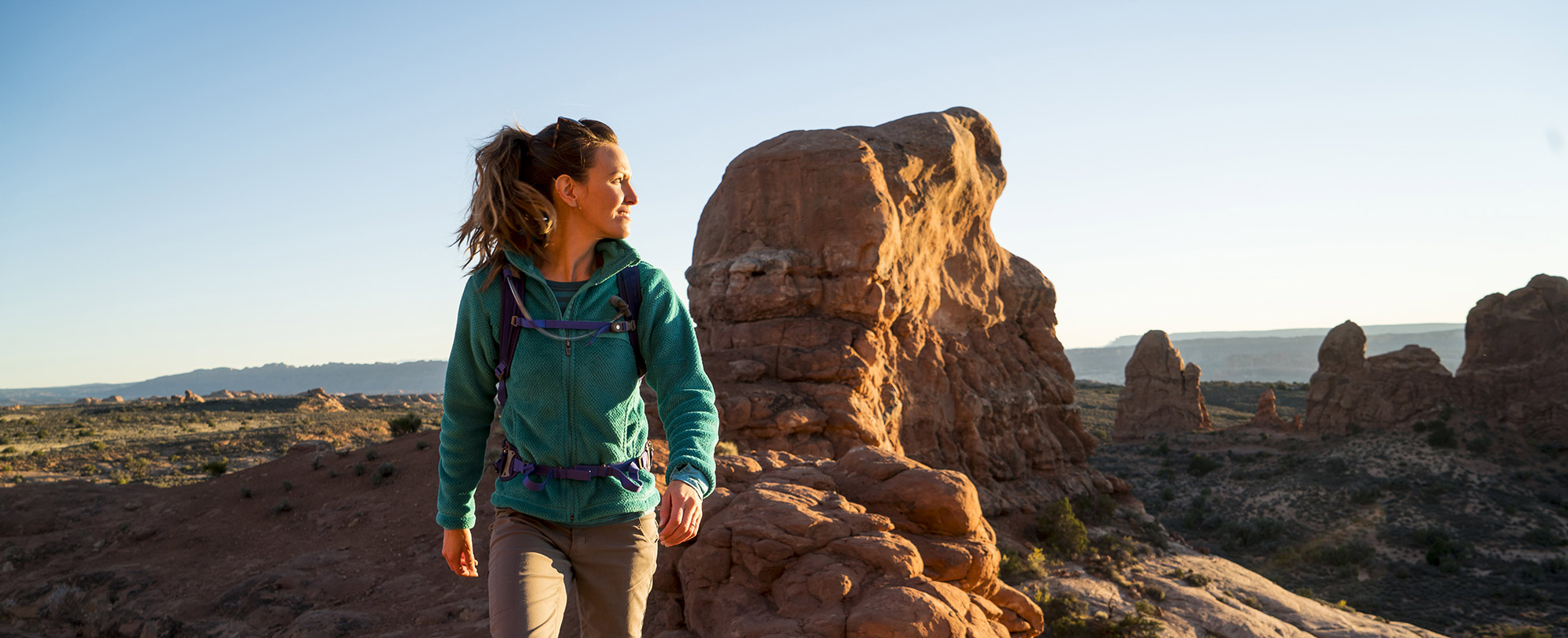 A woman in hiking gear looks back over her shoulder at large red rock formations while exploring the American Southwest.	