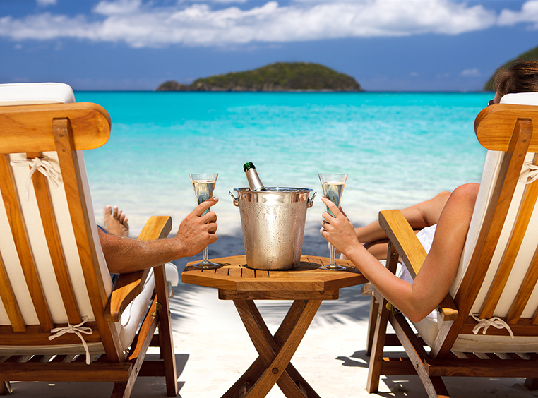 View of a man and a woman from behind as they lounge in beach chairs with a bottle on ice between them and each holding a glass of champagne as they look out at the ocean.