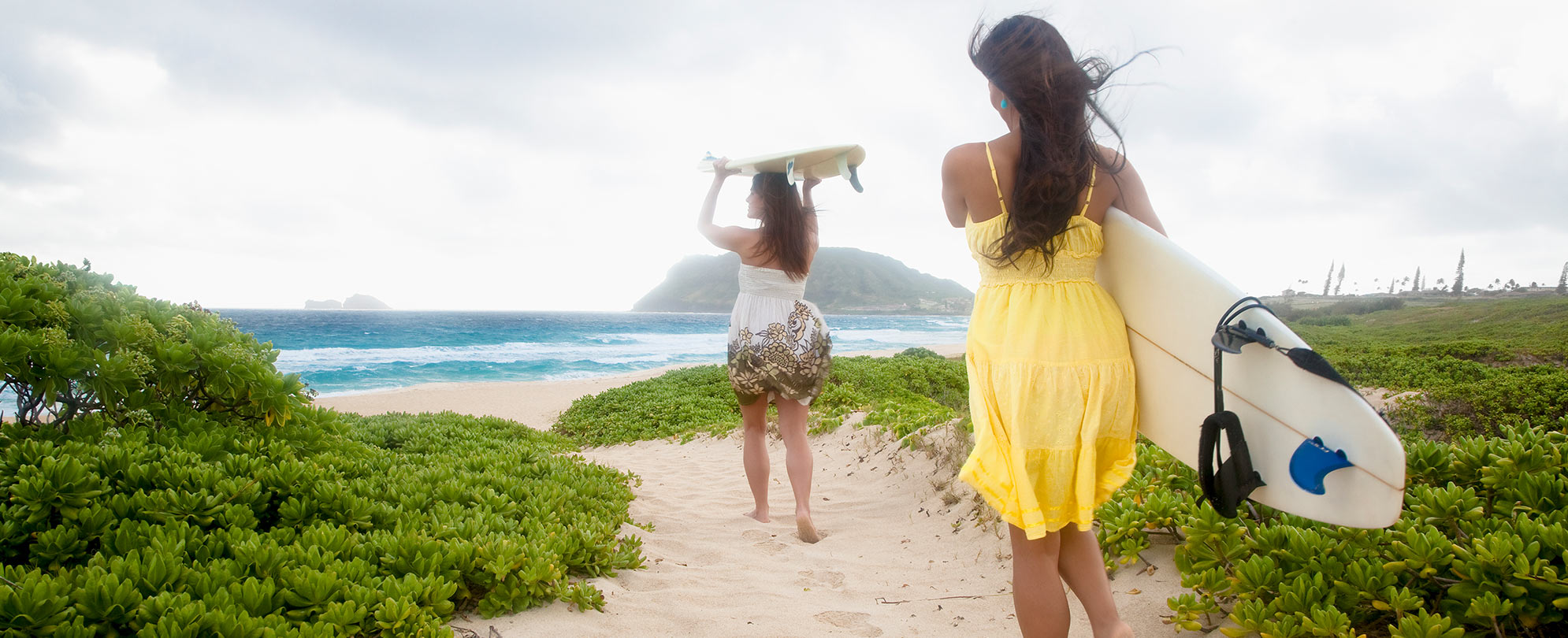 Two women surfers wearing sundresses carry surfboards down a sandy path towards the ocean in Hawaii.